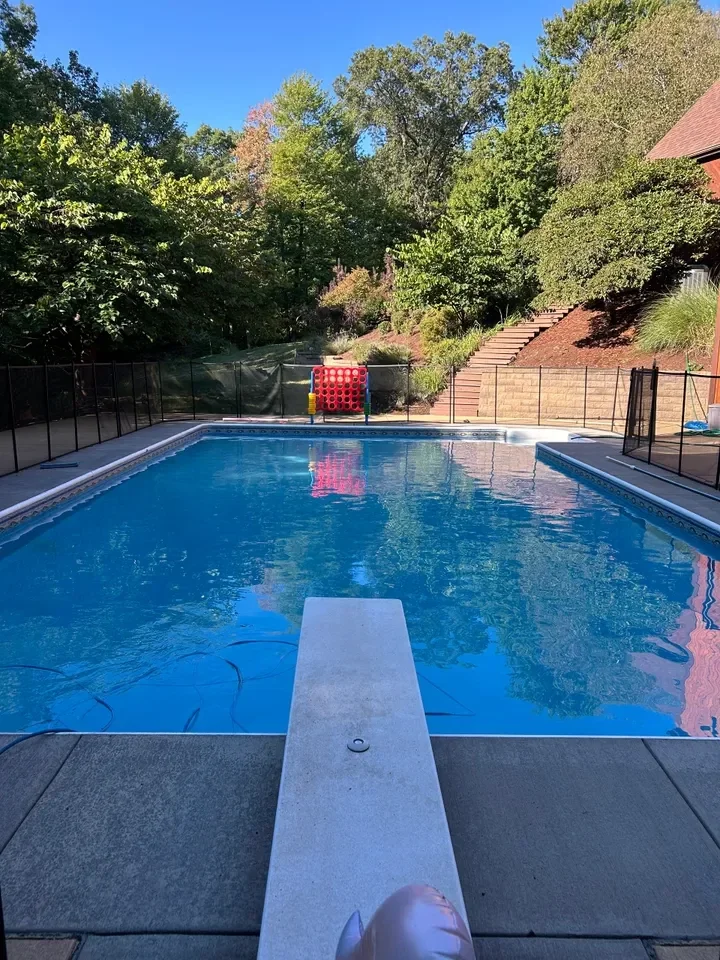 Residential swimming pool with diving board, surrounded by trees and brick structures on sunny day.