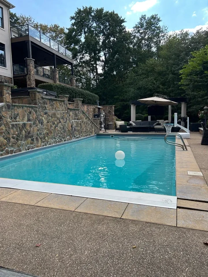 Modern backyard with turquoise swimming pool, stone walls, covered patio, and lush green trees