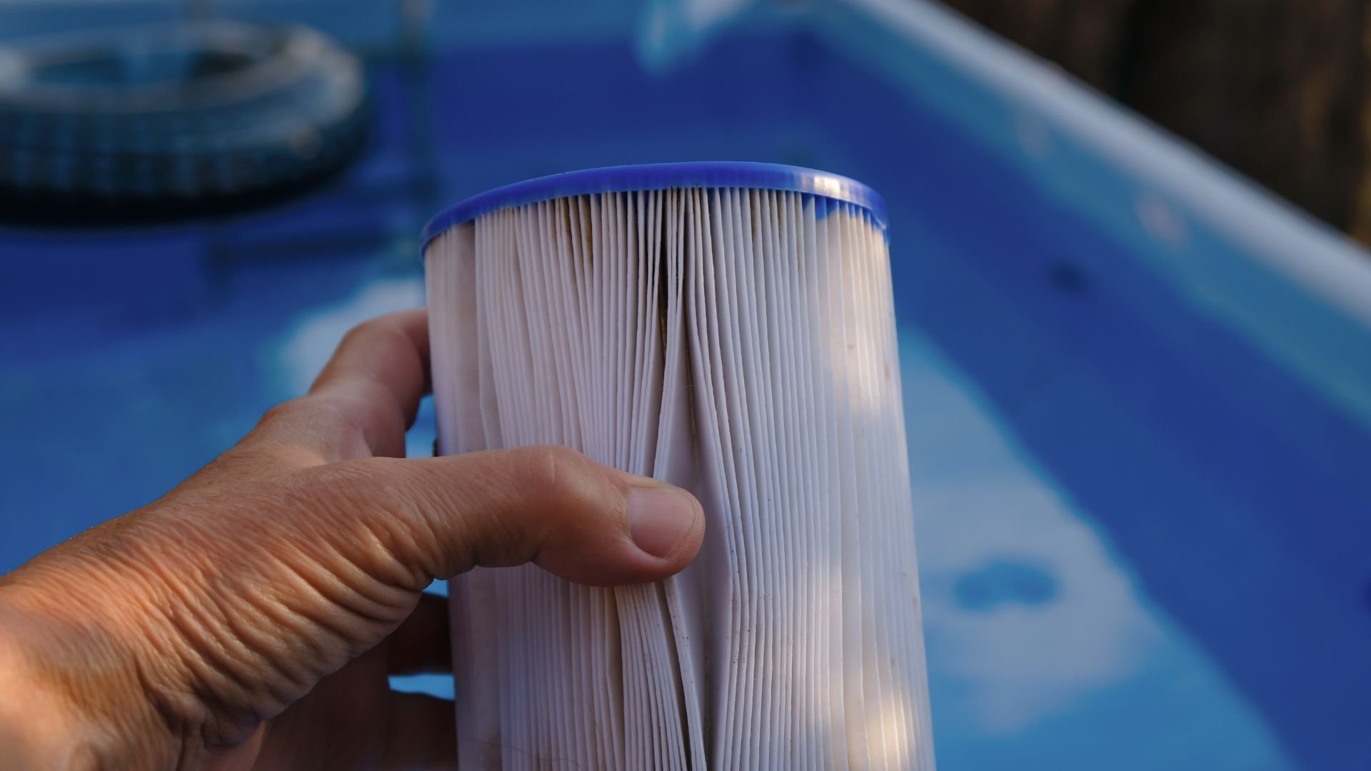 Hand holding a white pleated pool filter cartridge near blue water
