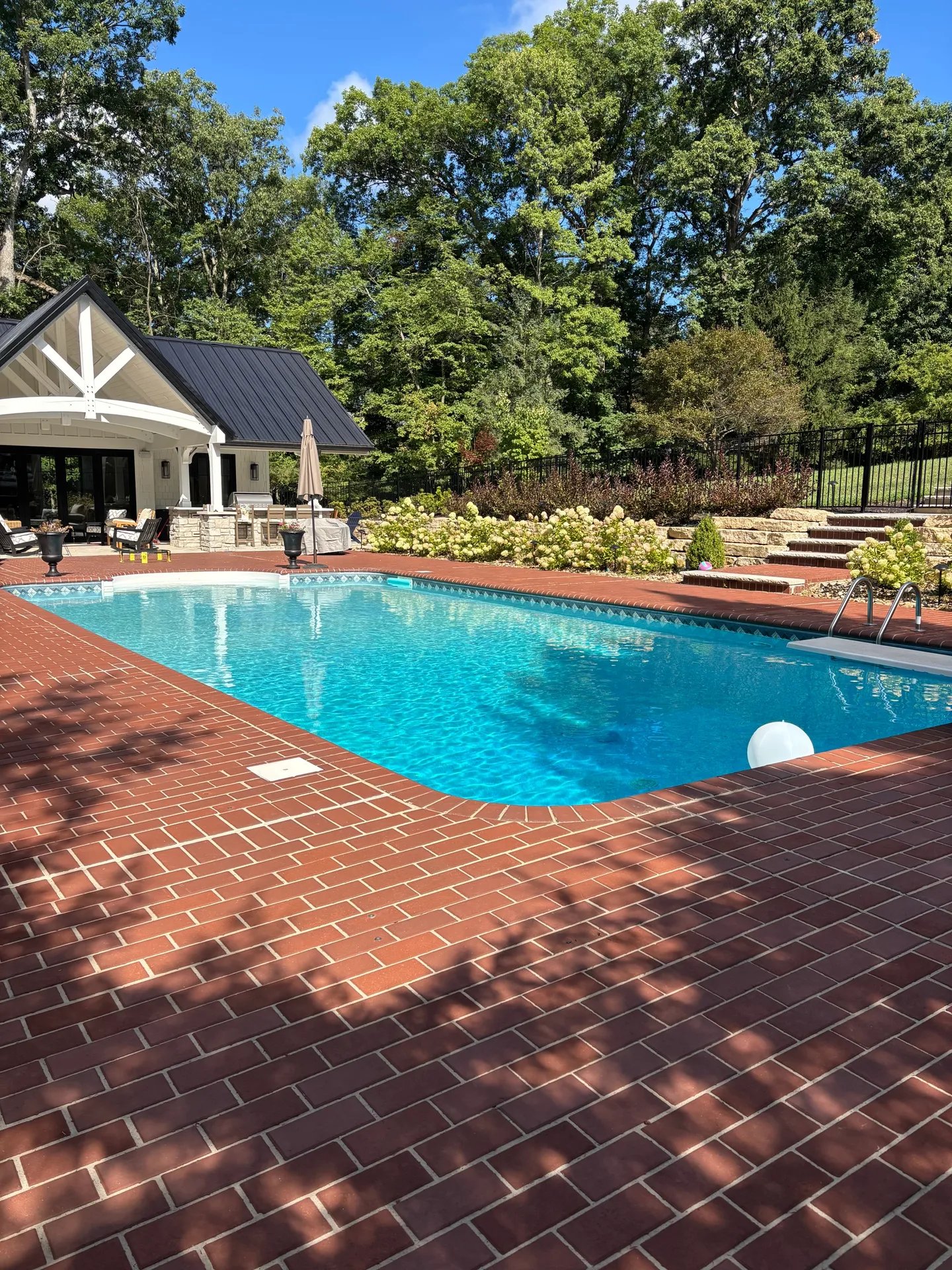 Brick-lined swimming pool with blue water near poolhouse and lush trees