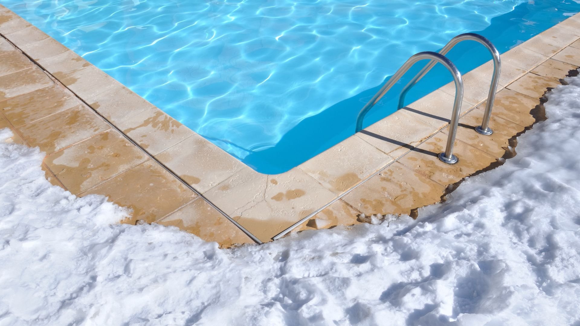 Swimming pool corner with bright blue water surrounded by snow and tiles