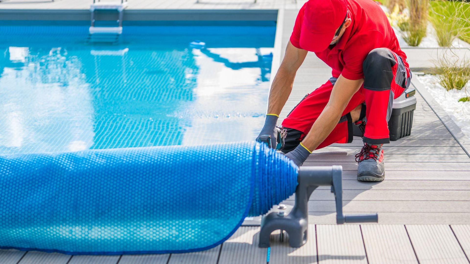 Pool maintenance worker in red rolling up blue solar cover near swimming pool